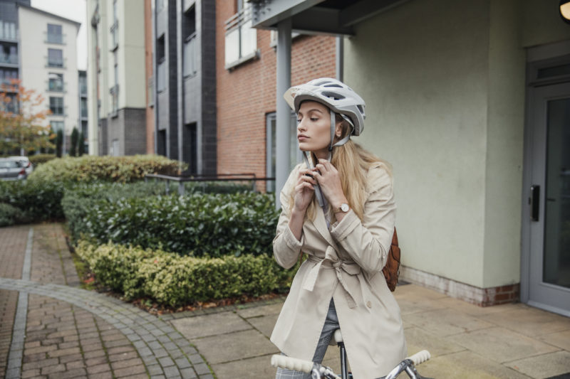 Attractive young woman putting on helmet as she sits on her bike directly outside her front door