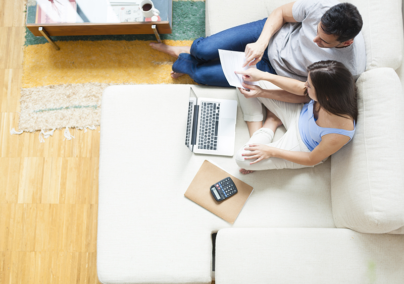 Aerial shot of couple on white couch looking at a piece of paper with a calculator, folder, and laptop alongside them