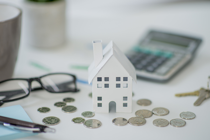 Tiny house made of paper alongside coins, coffee mug, coins, calculator, coin, and pen