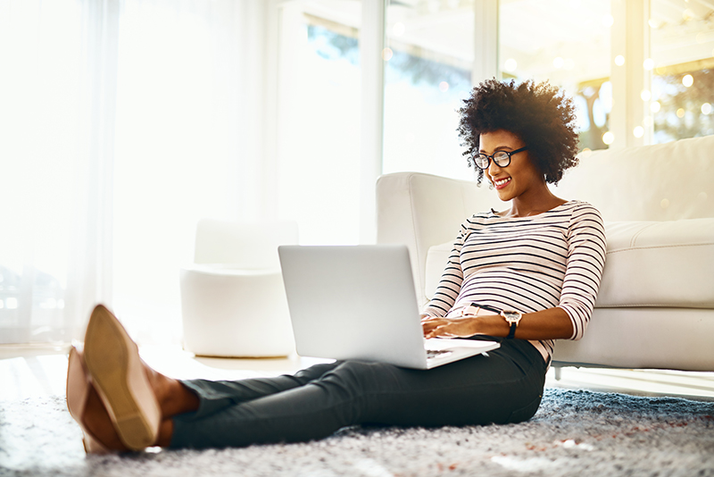 Young hip female smiling at her laptop as she sits on floor next to couch