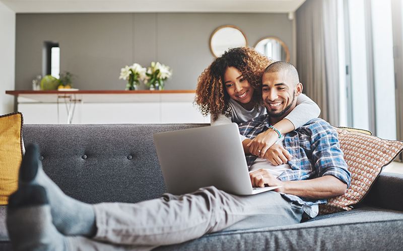 Young man smiling at his laptop with his girlfriend's arm wrapped around him on the couch