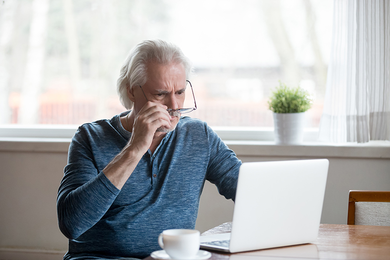 Elderly Male taking off glasses while looking at laptop screen at kitchen table